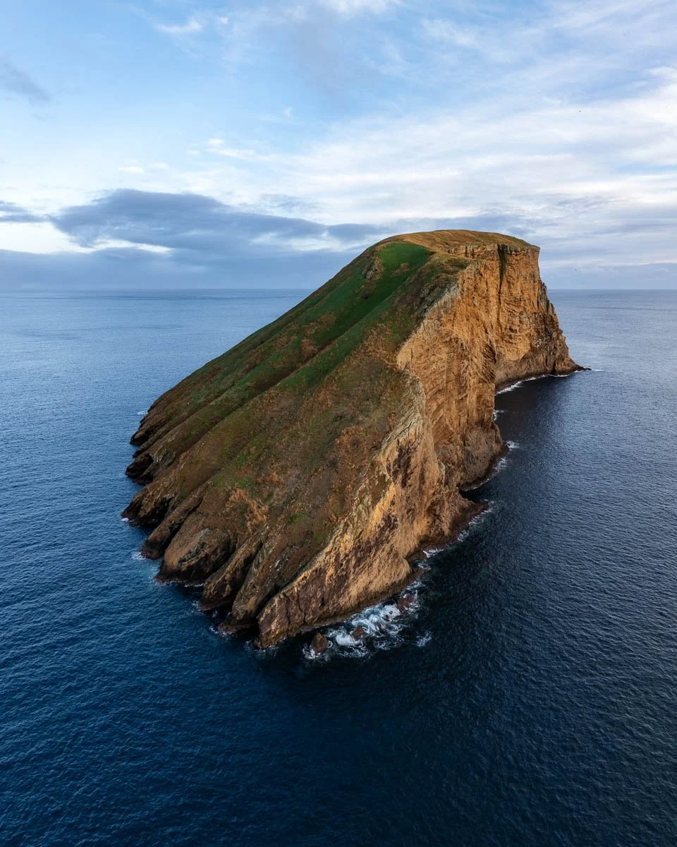 A large rocky island with grassy top, surrounded by the ocean under a sky with clouds.
