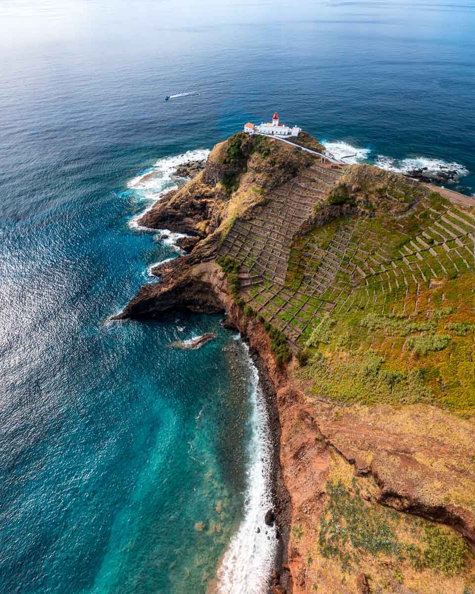 Aerial view of a lighthouse on a cliff overlooking the ocean, with terraced vineyards and greenery on the slope.