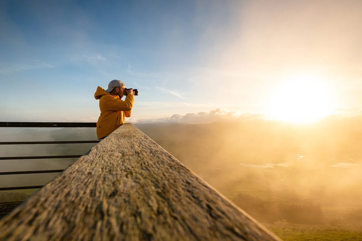 Person in yellow jacket and white cap taking a photograph from a mountain lookout during sunrise or sunset.