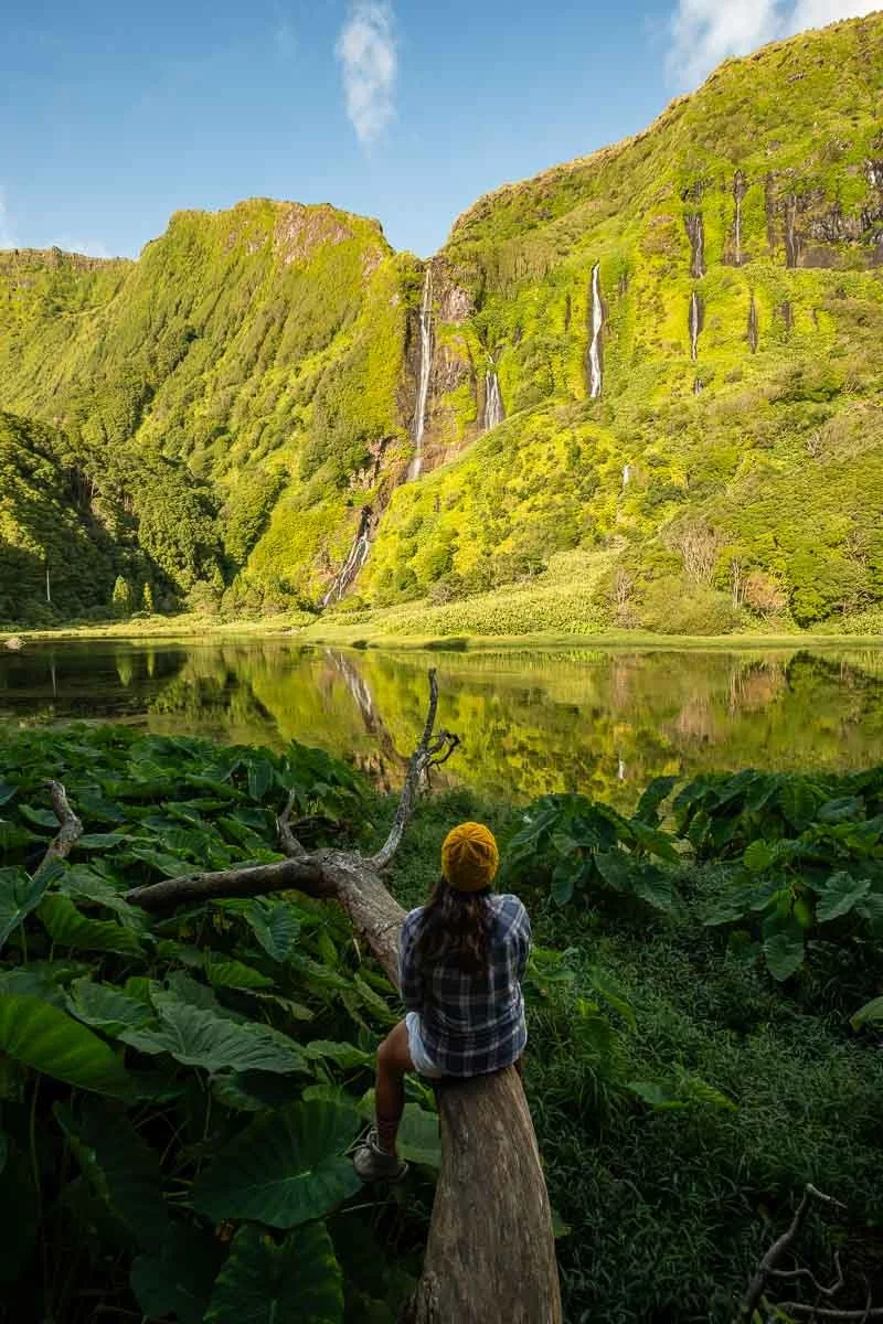 A woman with a yellow hat and plaid shirt sitting on a log near water, overlooking a lush green mountain landscape with waterfalls and a calm lake reflection.