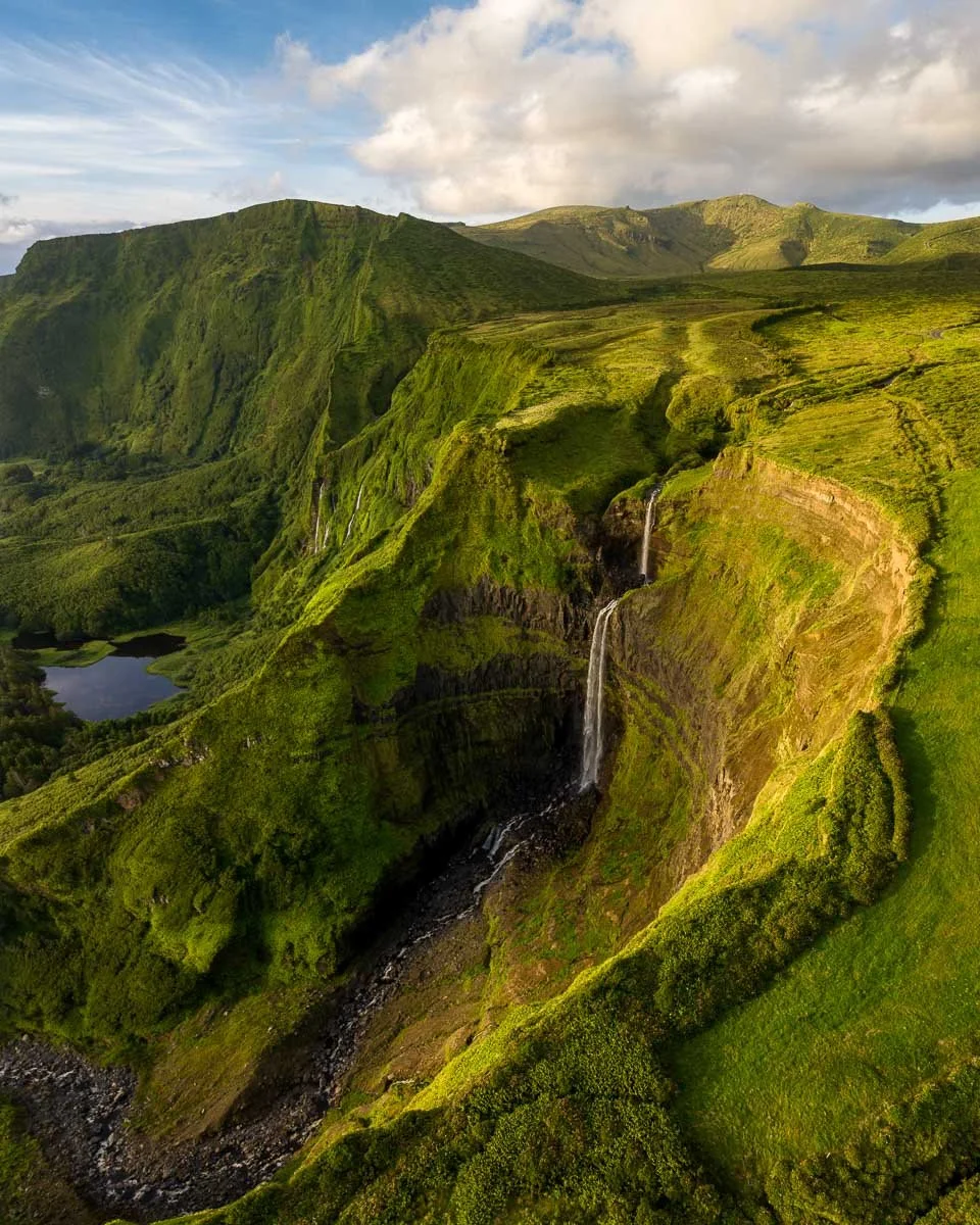 A lush, green landscape with mountains, waterfalls, and a small lake, taken during daylight with a partly cloudy sky.