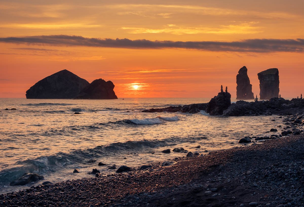 Sunset over a rocky beach with large sea stacks in the distance and waves crashing on the shore.