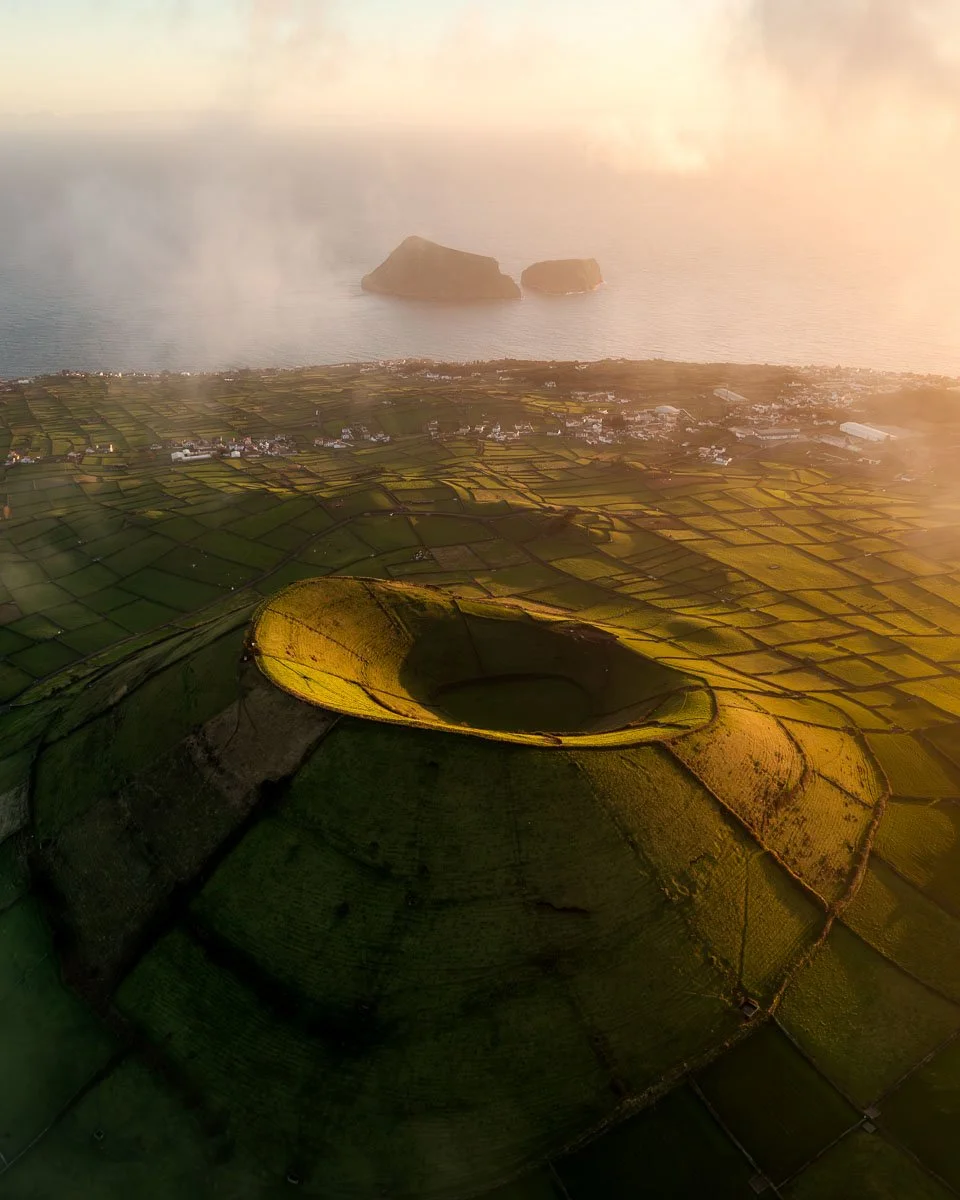Aerial view of a volcanic crater surrounded by green fields and overlooking the ocean with islands in the distance, illuminated by warm sunlight.