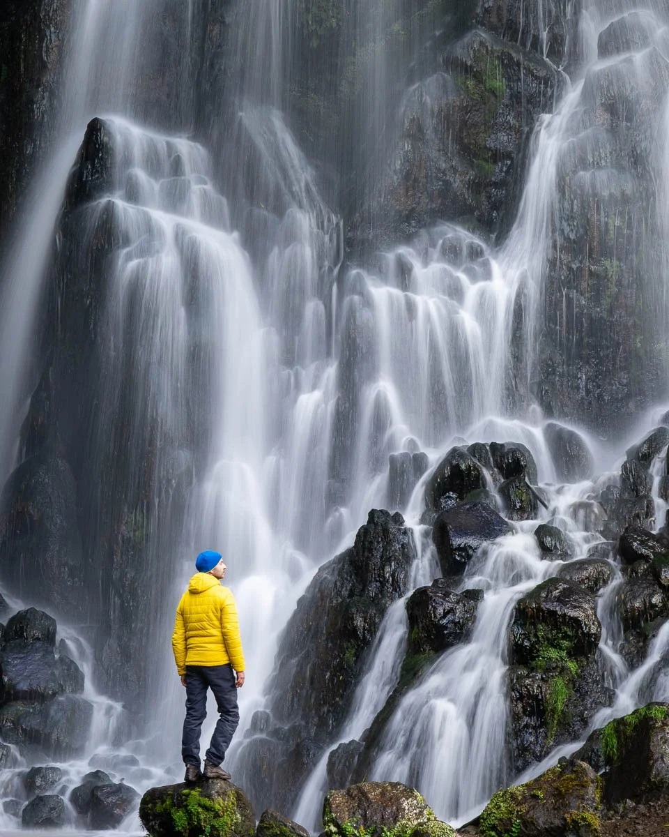 A person wearing a yellow jacket, blue beanie, and dark pants standing on mossy rocks in front of a large cascading waterfall.
