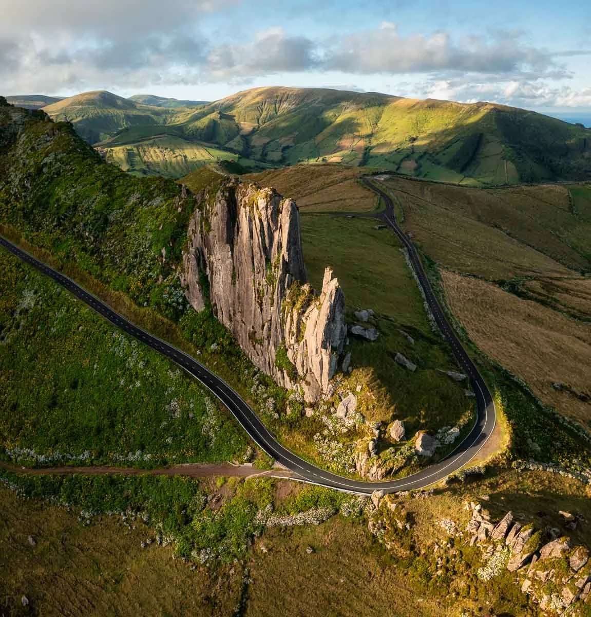 An aerial view of a winding road around a large, jagged rock formation in a green, hilly landscape with mountains in the background.