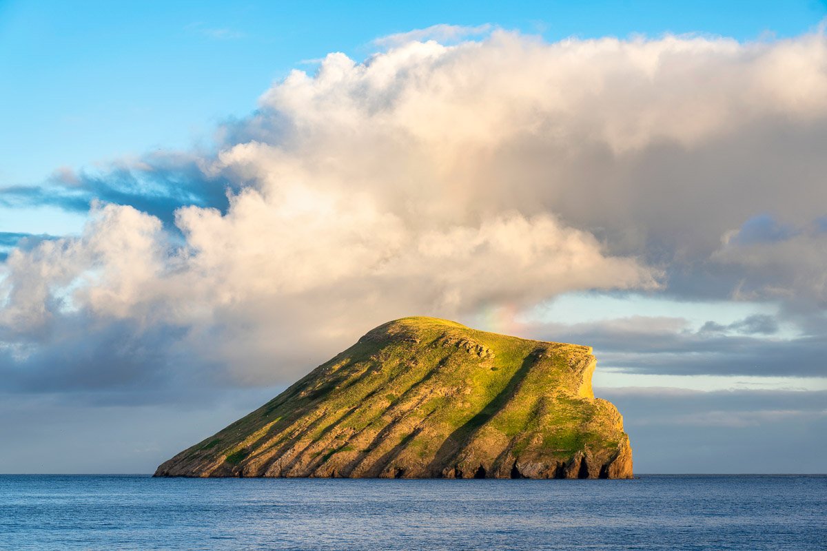 A large, steep, green-covered sea stack island in the ocean under a partly cloudy sky with a faint rainbow.