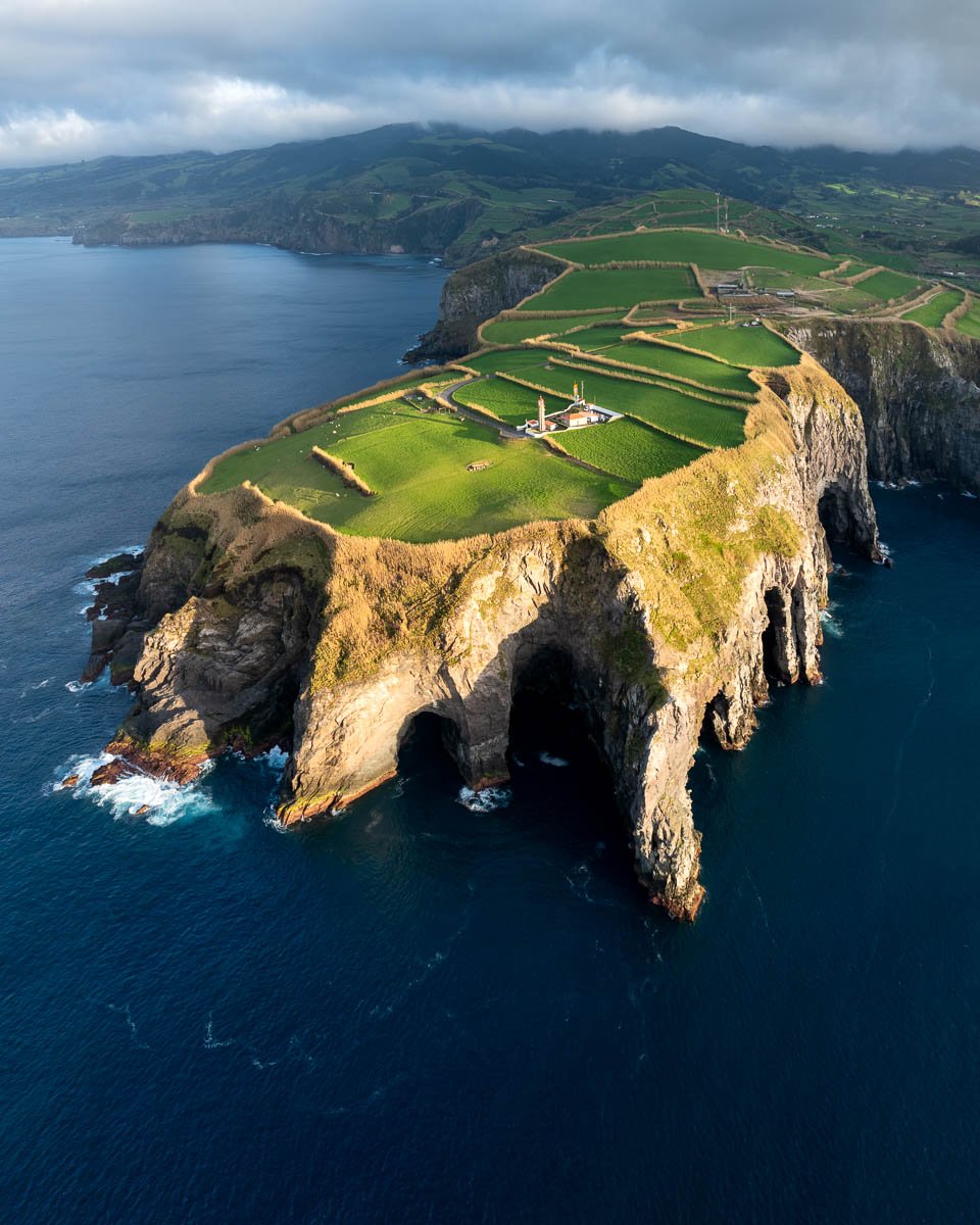 Aerial view of a large rocky island with green grass-covered cliffs featuring natural arches, surrounded by deep blue ocean waters, with rolling green hills and cloudy sky in the background.