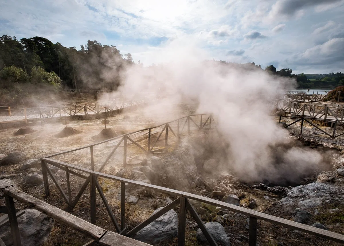 Geothermal area with steam rising from hot ground, wooden walkways, and rocky terrain under a cloudy sky.