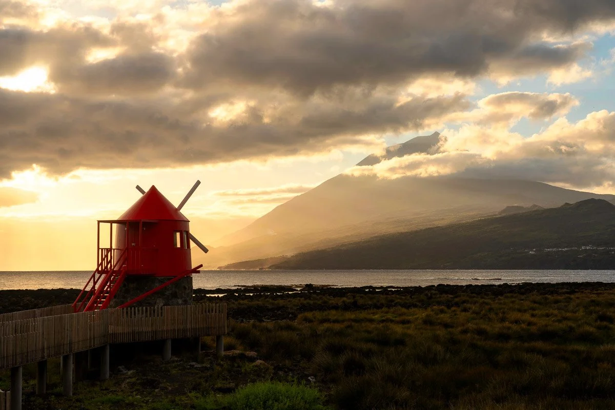 A small red windmill on a stone base with a wooden walkway leading to it, set against a landscape with a body of water and a mountain in the background during a cloudy sunset.