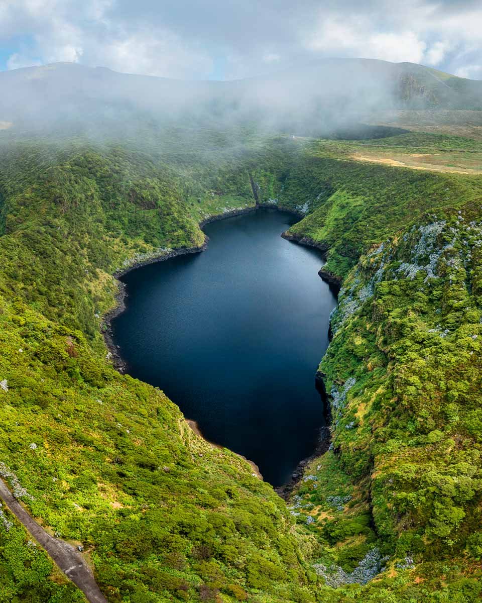 A deep, dark water-filled crater surrounded by lush green vegetation and misty mountains in the background.