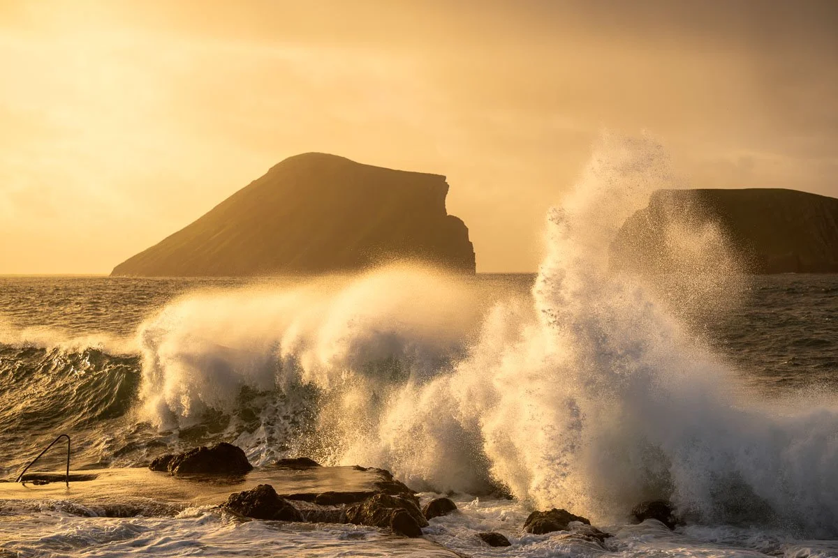 Waves crashing against rocks in the ocean during sunset with two large rocky islands in the background