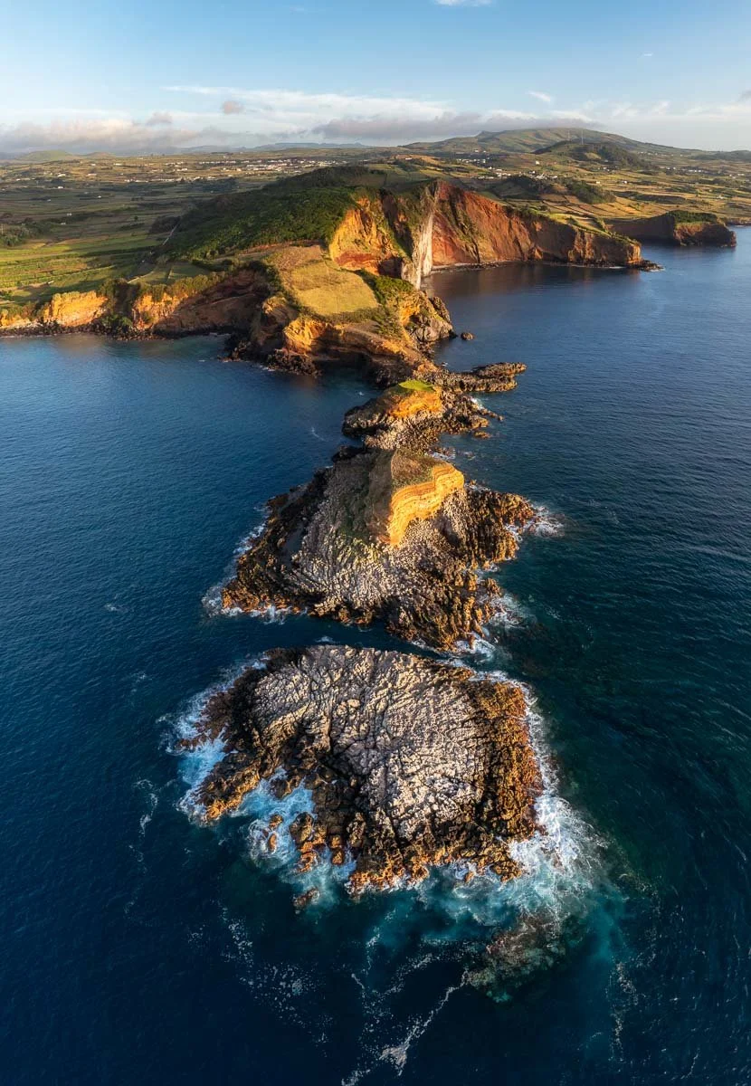 Aerial view of rocky coastline with cliffs and ocean, green hills and farmland in the background.