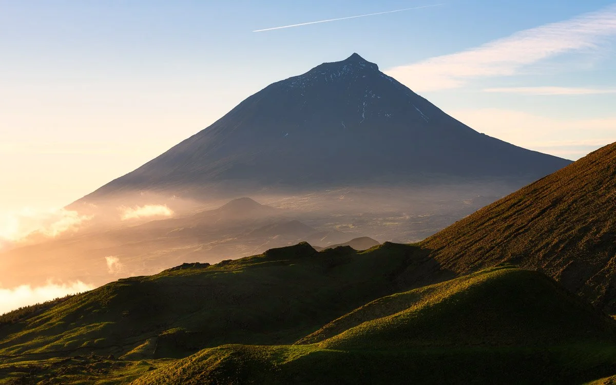 A mountain with a conical shape, likely a volcano, with a snow-capped peak and grassy slopes in the foreground, during sunrise or sunset.