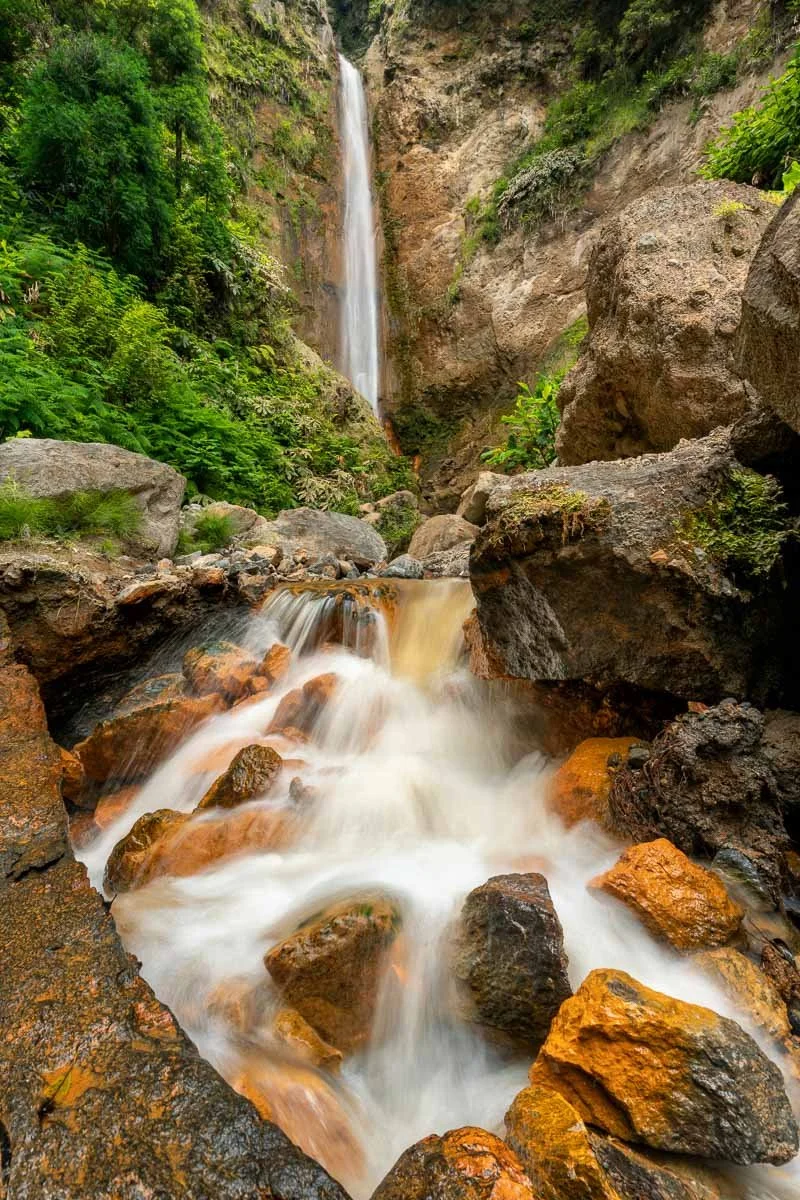 A waterfall flowing down a rocky cliff into a stream surrounded by lush green vegetation.