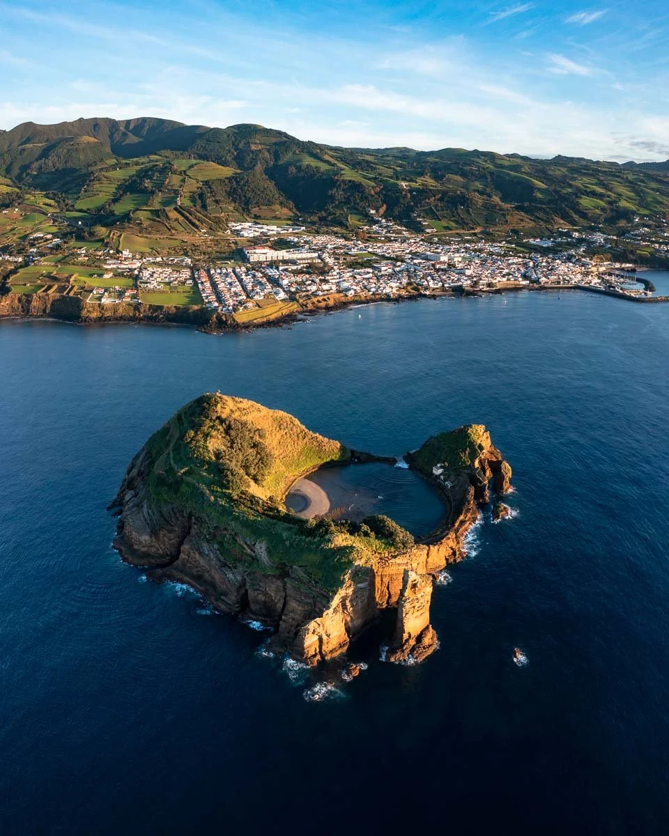 Aerial view of a coastal town with a hilltop harbor surrounded by ocean and green mountains in the background.