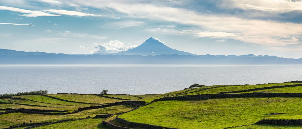 Green rolling hills with stone walls near a body of water, mountain with snow-capped peak in the background, partly cloudy sky.