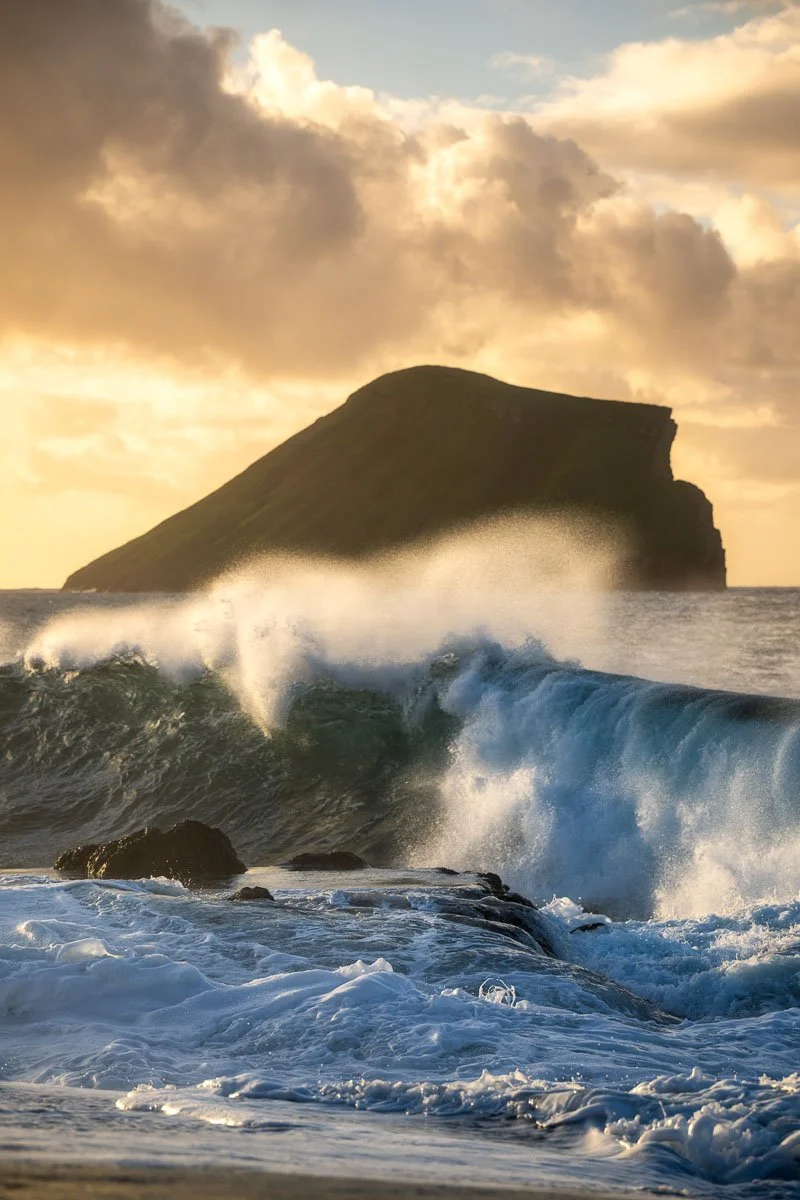 Ocean waves crashing against rocks with a large, rounded island or mountain in the background during sunset or sunrise with cloudy sky.