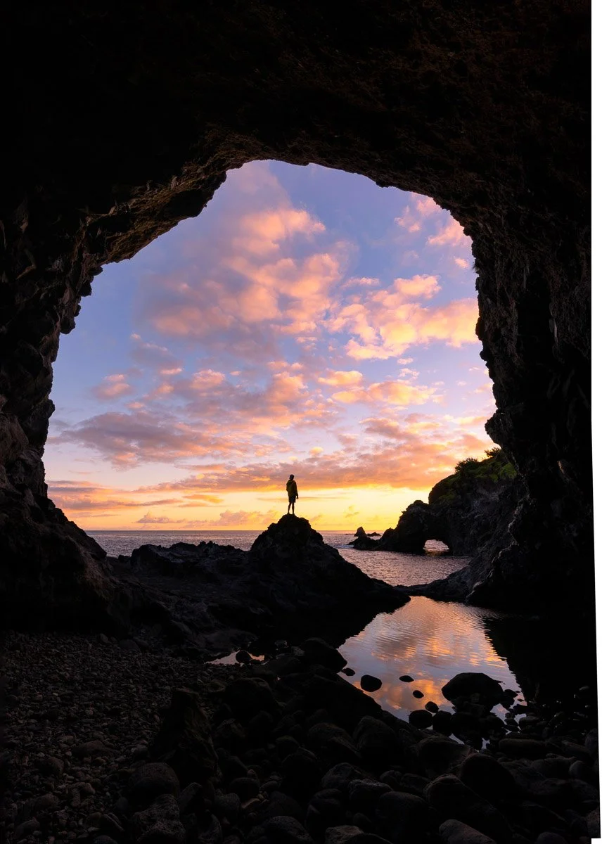 A person stands atop rocks at the entrance of a cave, with a colorful sunset sky and ocean in the background, and reflections on the water below.