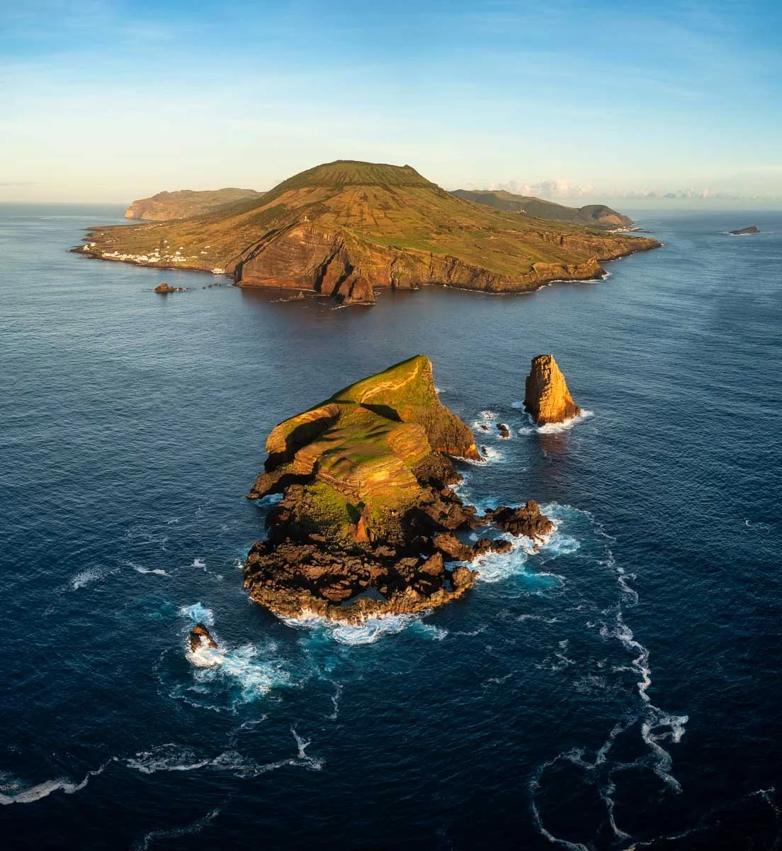 Aerial view of a volcanic island with steep cliffs, green fields, and surrounding ocean with waves crashing against rocks.