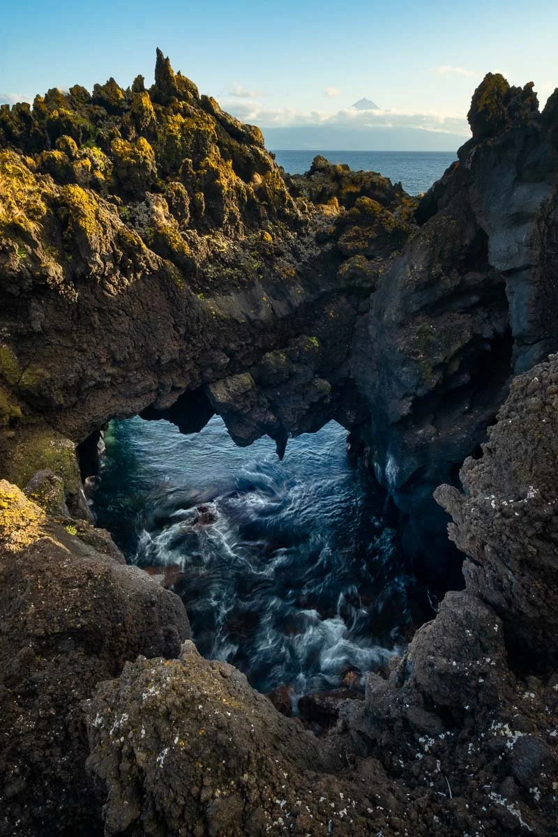 Coastal rock formations with a small sea arch and ocean water beneath, with a distant mountain or volcano on the horizon under a clear sky.