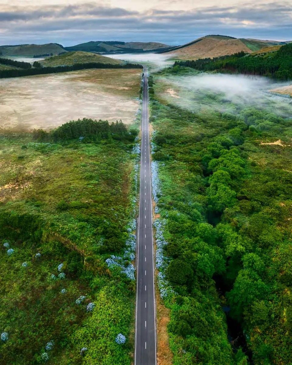 An aerial view of a long, straight road running through lush green countryside with hills in the background, some fog and clouds in the sky.