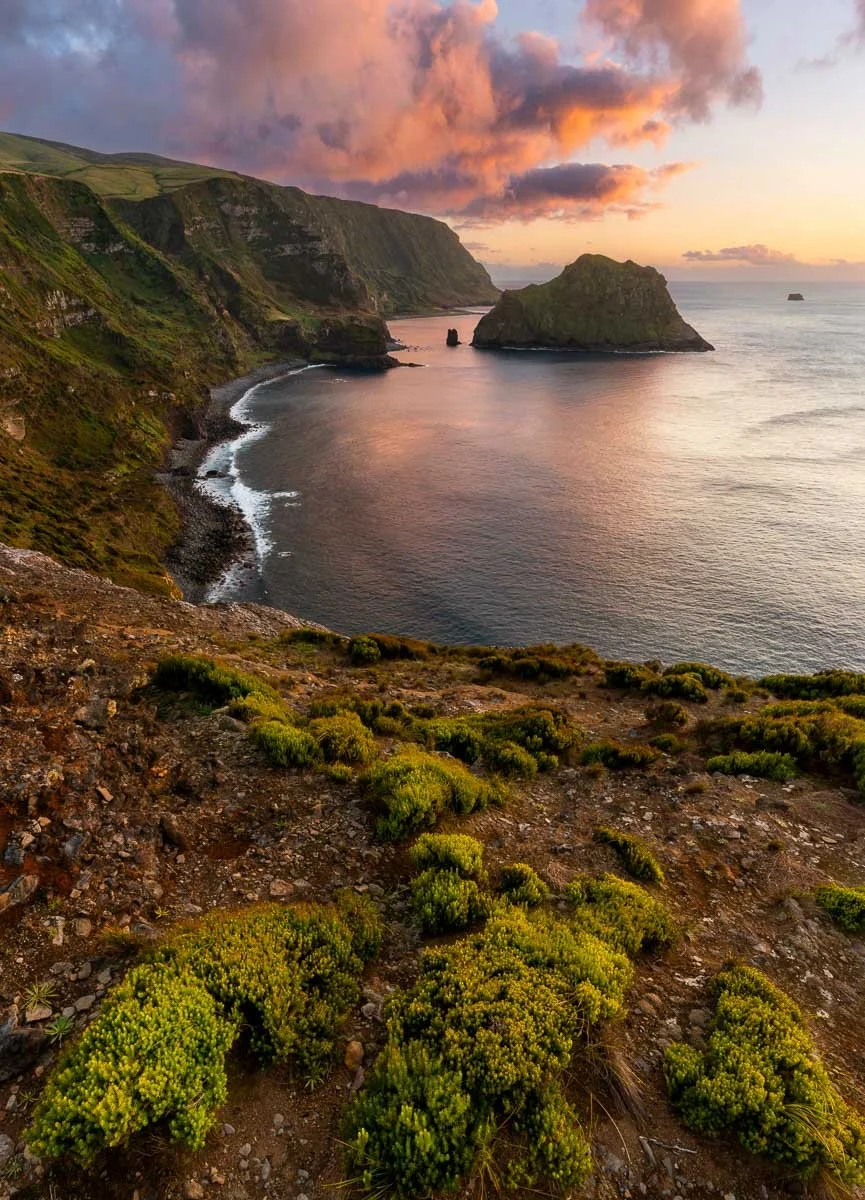 Scenic coastal landscape with green cliffs, a large sea stack, and a vibrant sunset sky with pink and orange clouds over the ocean.