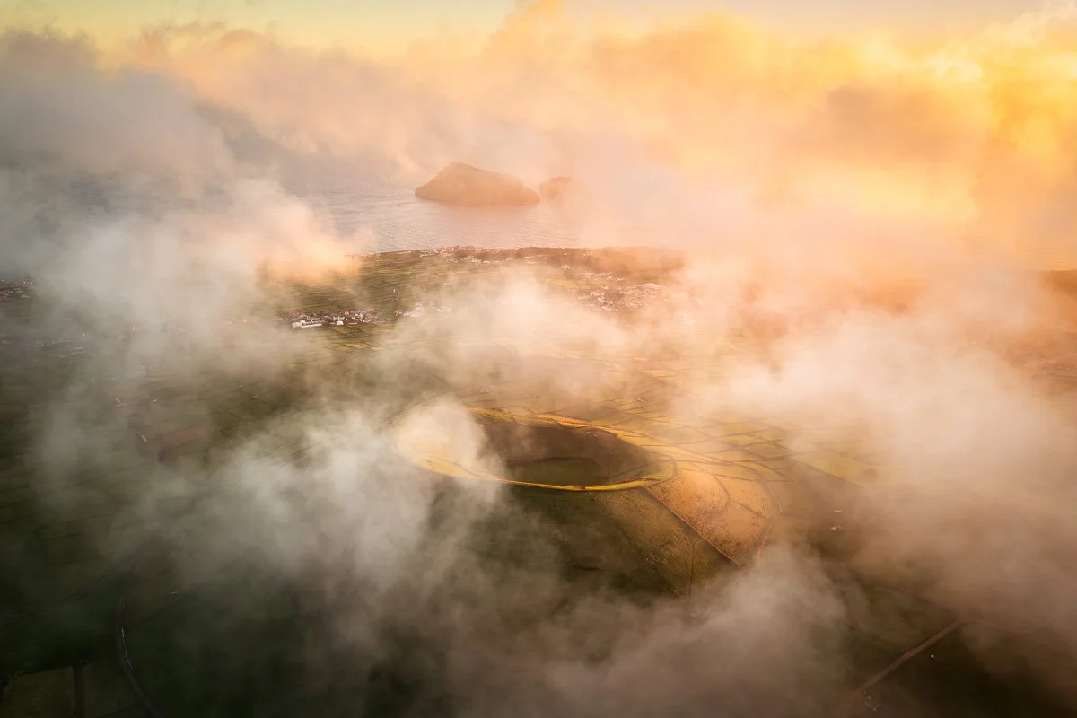 A landscape with a large volcanic crater in the foreground, surrounded by clouds of smoke and fog, and a body of water with a rocky island in the background.