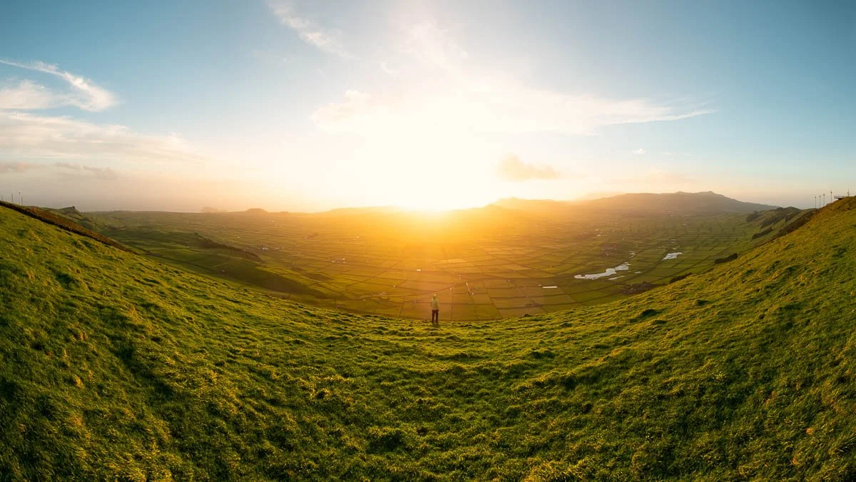 A person standing on a grassy hilltop overlooking a farmland valley at sunset with rolling hills and cloudy sky.