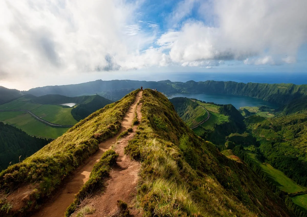A mountain trail on a green ridge overlooking a forested valley, a lake, and the ocean in the distance under a partly cloudy sky.