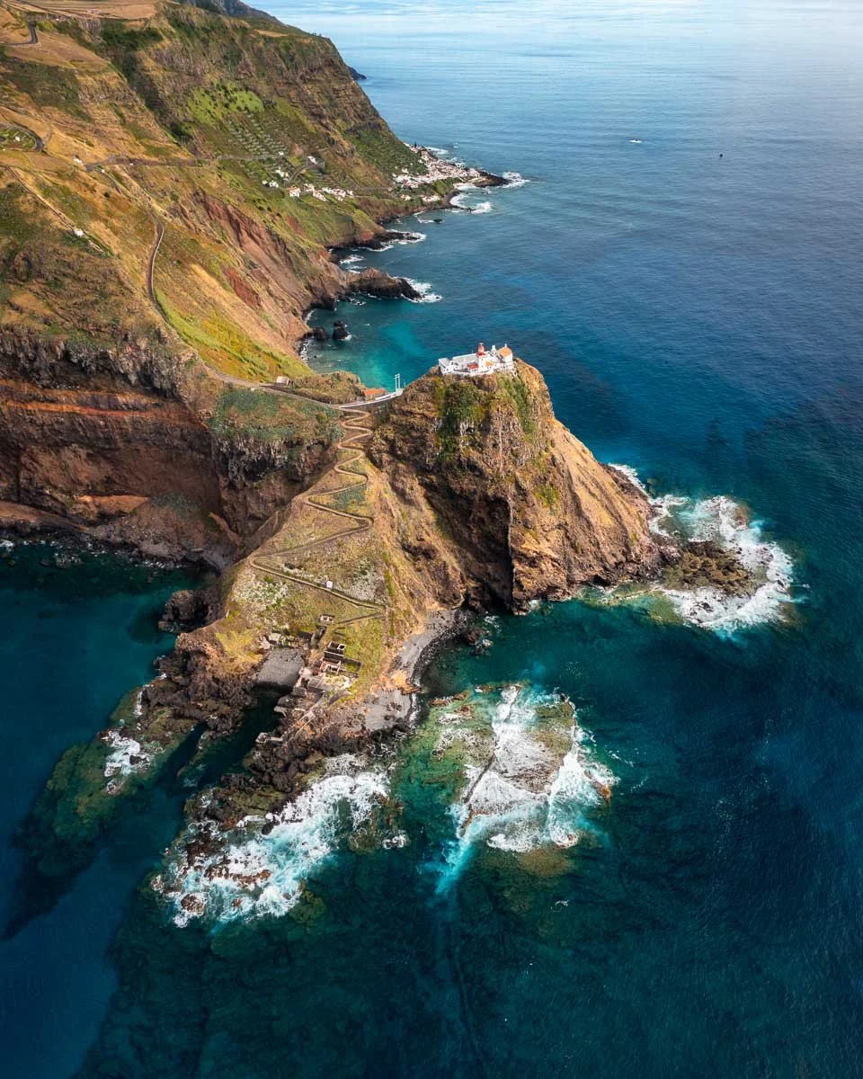 Aerial view of a rugged coastline with cliffs, a winding trail, and a white building on a small rocky promontory overlooking the ocean.