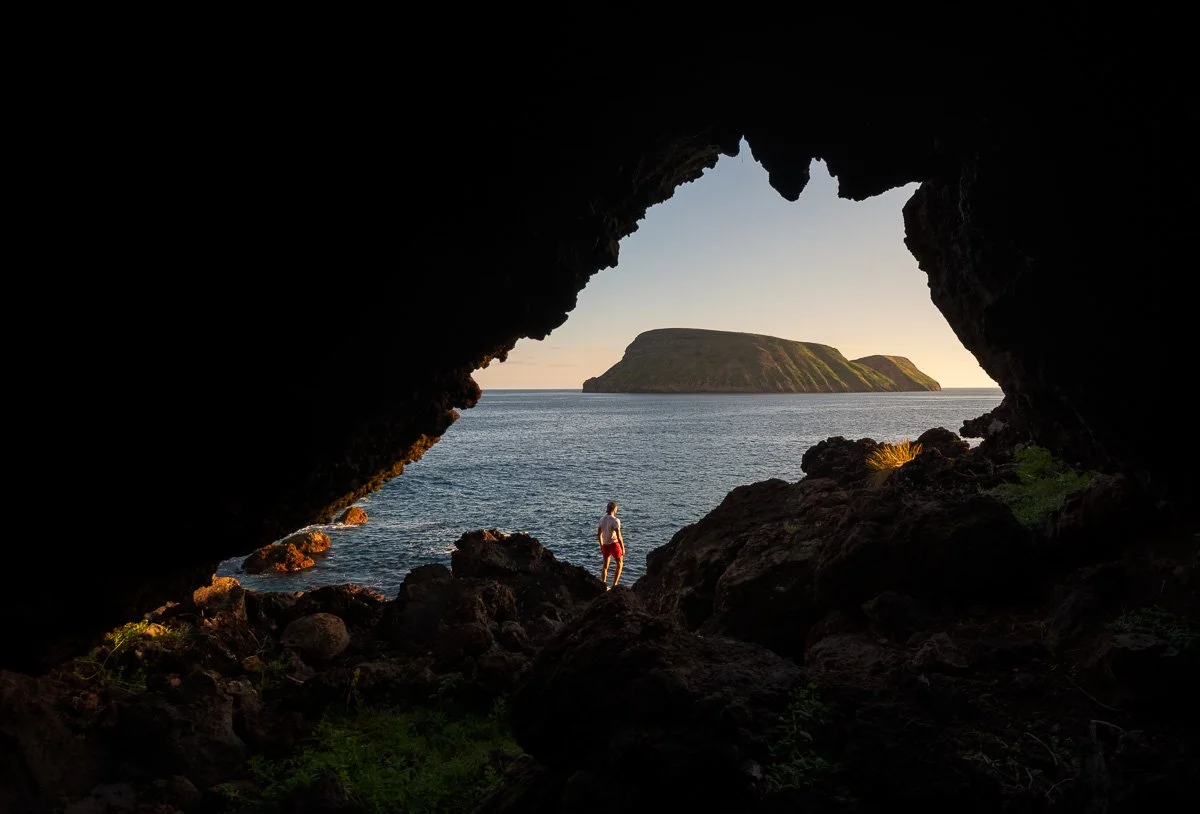 A person standing on rocky shoreline, viewed from inside a dark cave opening, with ocean and island visible at sunset.