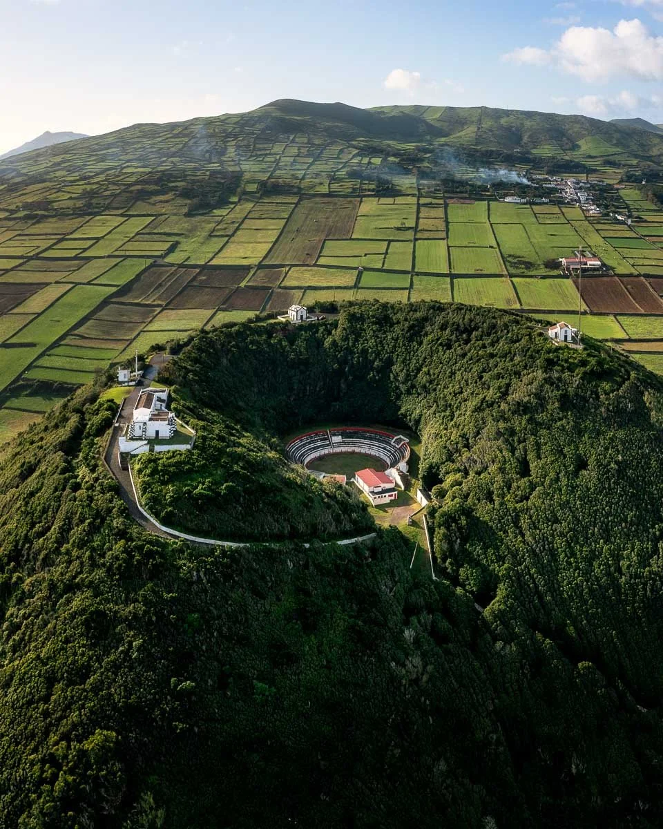 A lush green hillside with a circular amphitheater at the bottom, surrounded by dense trees, with several white buildings nearby and farmland in the background under a partly cloudy sky.