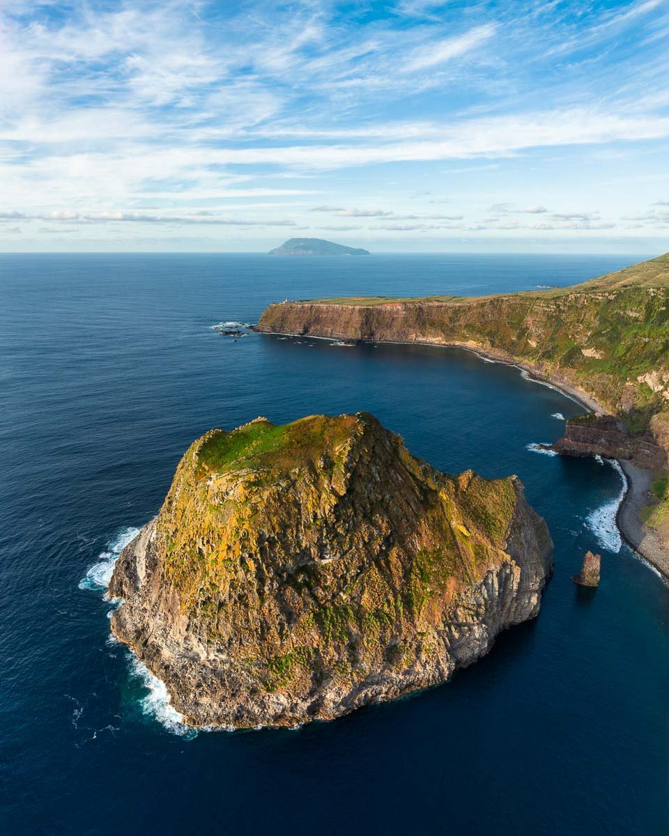 Aerial view of rugged coastal cliffs and a large rock formation surrounded by deep blue ocean with an island visible in the distance under a partly cloudy sky.