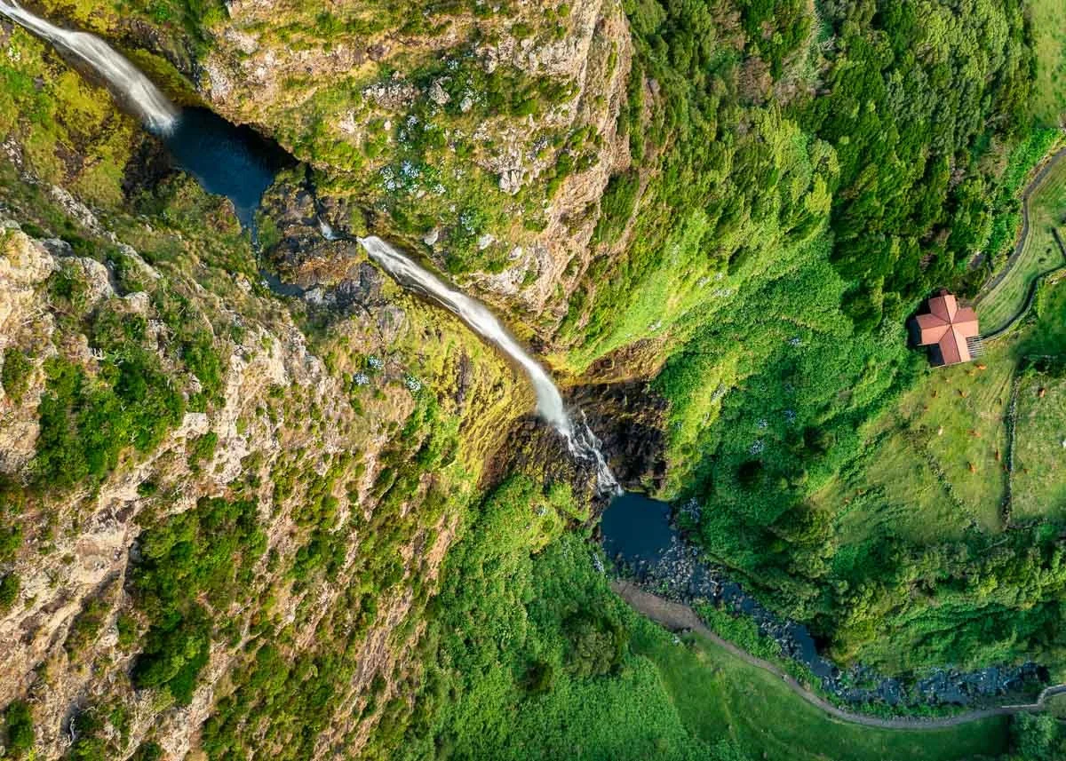 Aerial view of a waterfall flowing down a lush green canyon into a river, with a small house on the edge of the canyon surrounded by trees and grass.