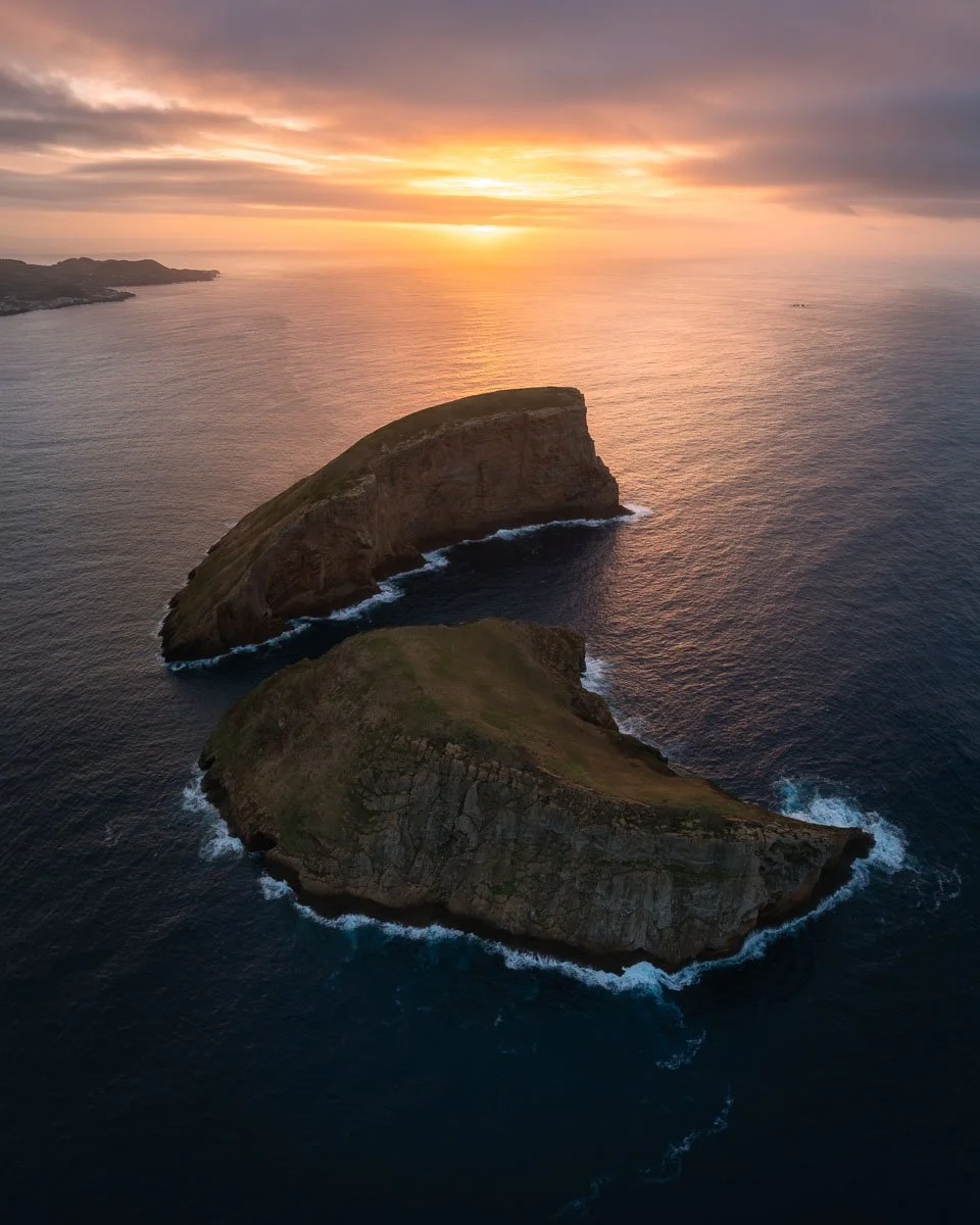 Aerial view of two large, rocky islands surrounded by ocean at sunset, with a colorful sky and distant coastline.