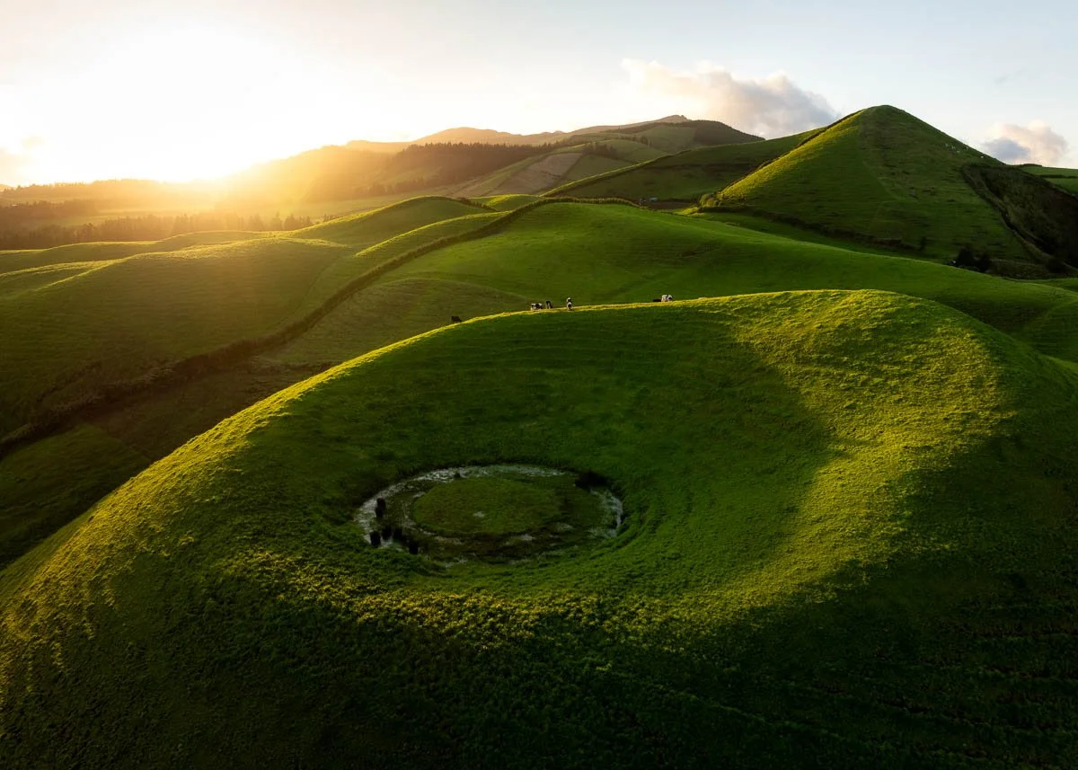 Sunset over green rolling hills with small circular water feature in the foreground.