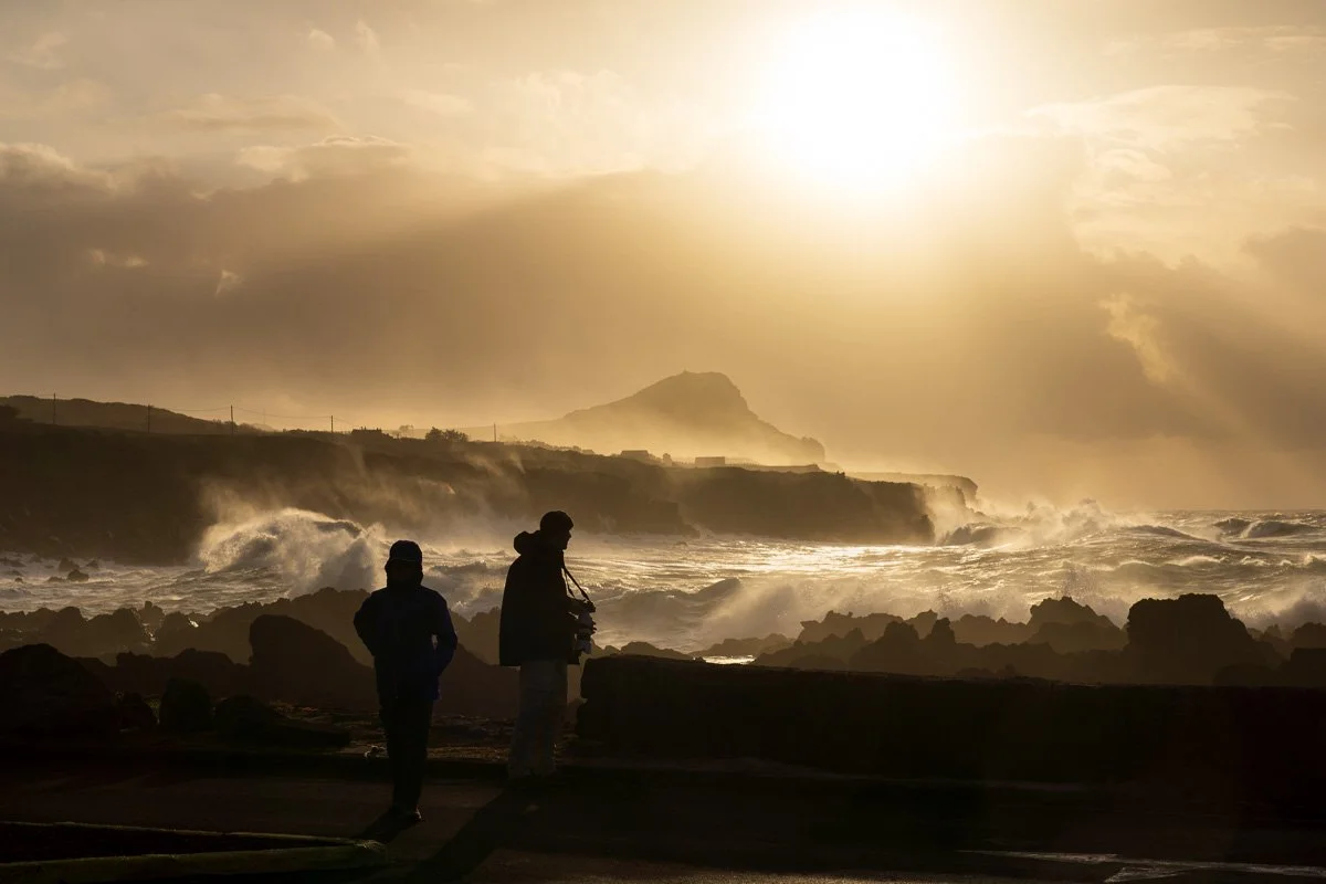 Silhouettes of two people standing on a rocky coastline during sunset with large waves crashing and a mountain in the background.