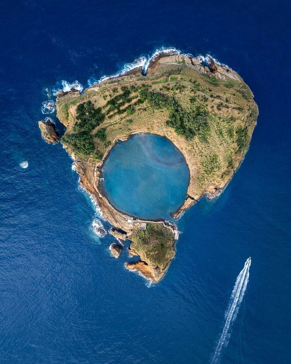 Aerial view of a small island with a circular lagoon surrounded by rocks, greenery, and ocean, with a boat leaving a wake in the water.