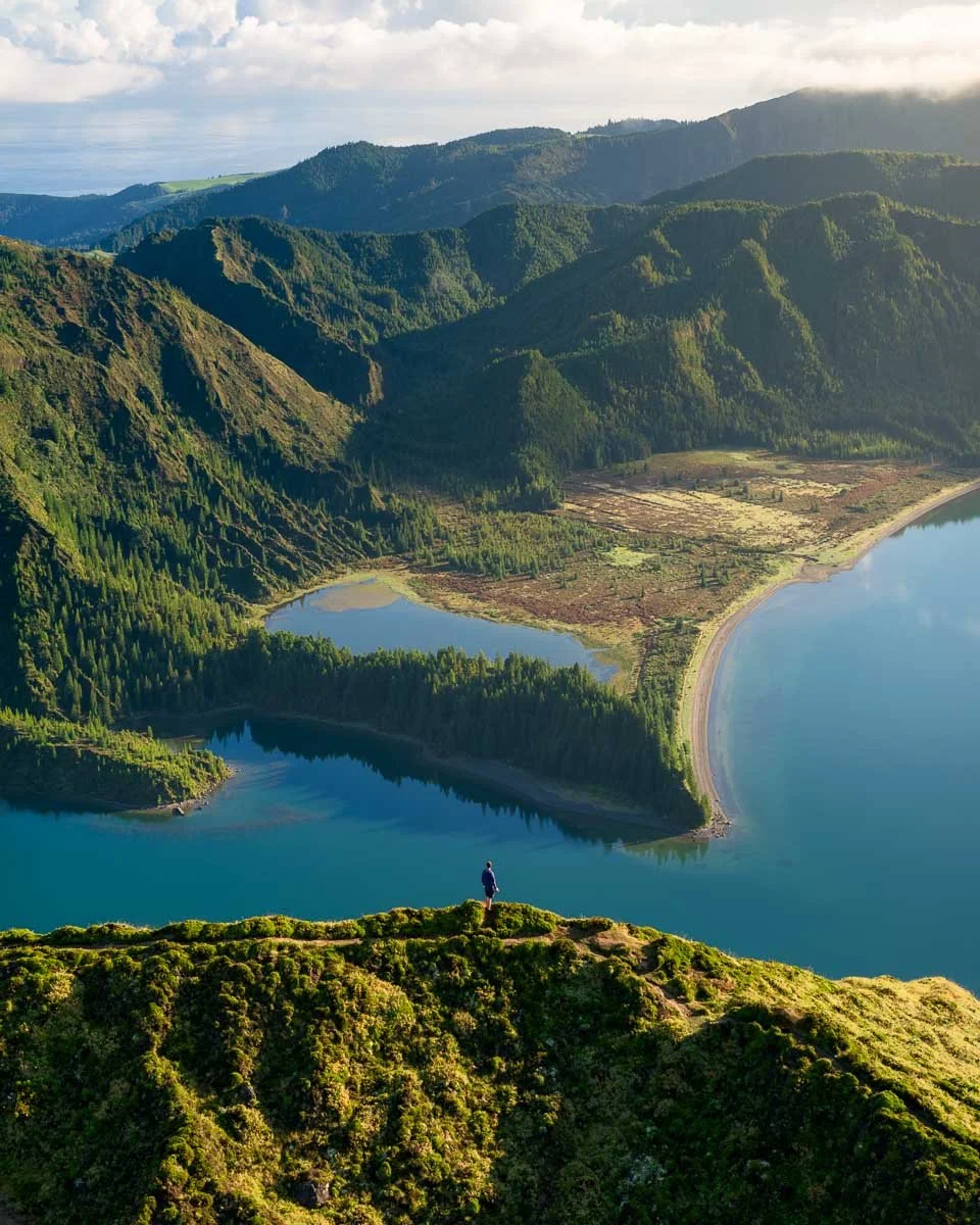 A person standing on a grassy hill overlooking a large body of water, surrounded by lush green mountains with dense forests and a partly cloudy sky.