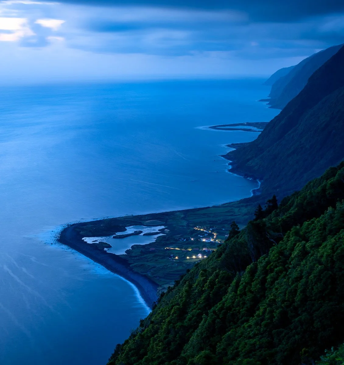 A coastal landscape at dusk with a steep, green mountainside on the right, a village with illuminated houses at the base, and the calm ocean extending to the horizon under a cloudy sky.