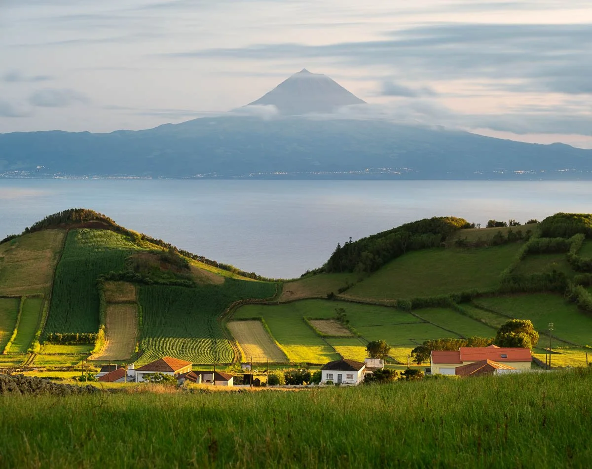Scenic landscape with green fields and small houses in the foreground, a large lake in the middle, and a mountain in the background, likely Mount Fuji.