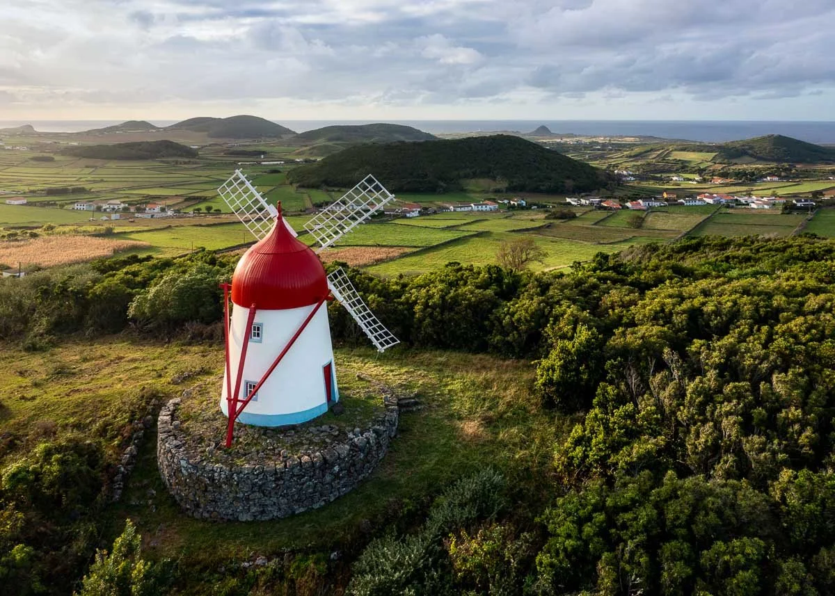 A windmill with a red roof and white body, situated on a stone base, surrounded by green bushes and fields, with hills and the ocean in the background.