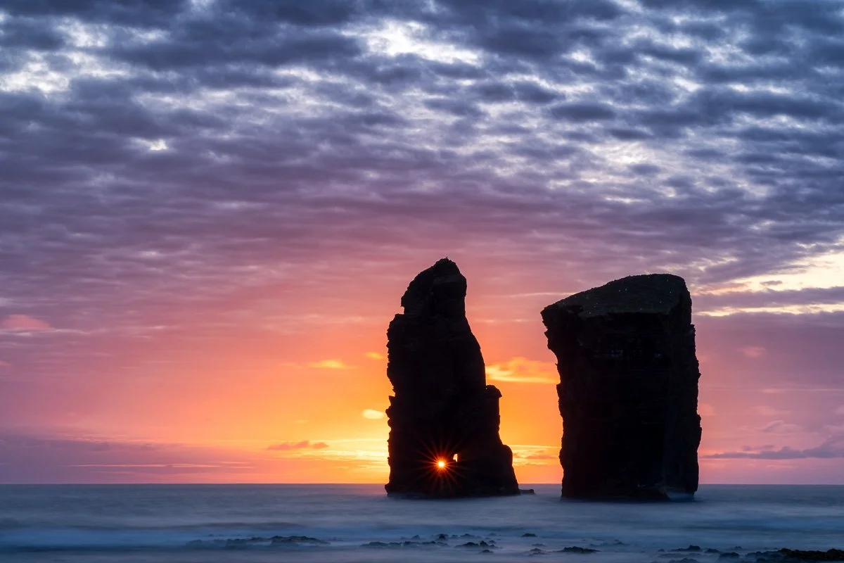 Sunset over the ocean with two large rock formations in the water. The sun is partially visible through a gap in the left rock, creating a starburst effect. The sky features purple and orange hues with scattered clouds.
