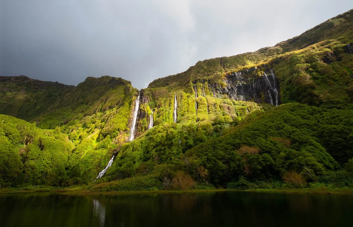 A lush green mountain landscape with several waterfalls flowing down the cliffs into a calm body of water at the base.