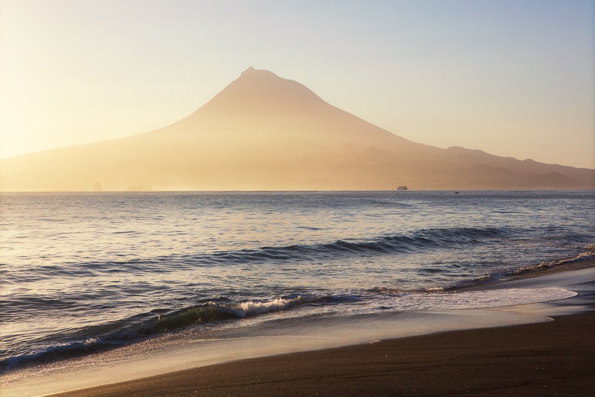 A sunset view of a mountain, possibly Mount Fuji, seen across a body of water from a beach with gentle waves.