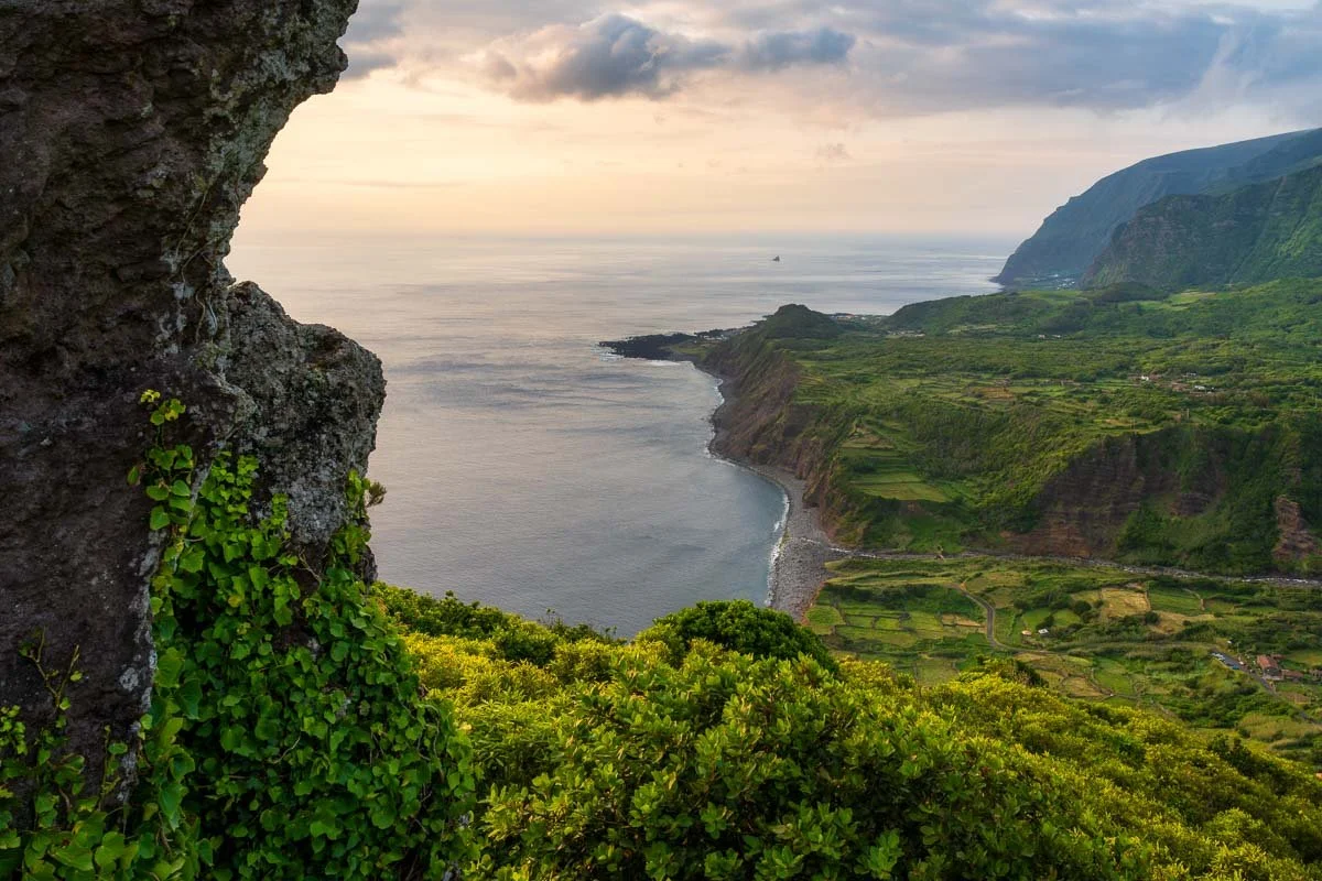 A scenic view of the coastline with green fields, cliffs, and ocean, seen from a vantage point with a rocky foreground and cloudy sky.