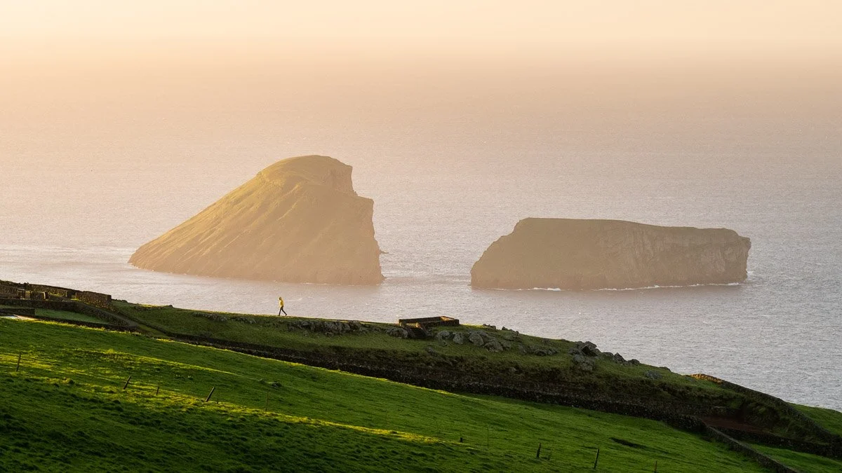 A person walking on a lush green grassy hillside near the coast, with two large rocky sea stacks in the ocean during sunset or sunrise.