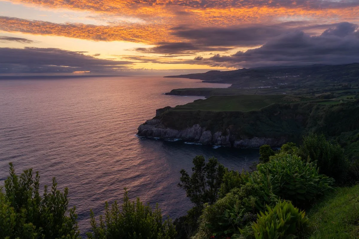 Sunset over a coastal landscape with cliffs, green hills, and the ocean, with partly cloudy sky.