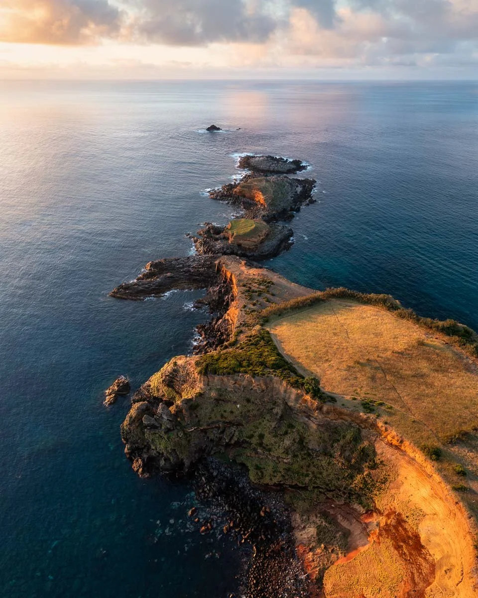 Aerial view of a rugged coastline with rocky outcrops and cliffs extending into the ocean, lit by sunset light.