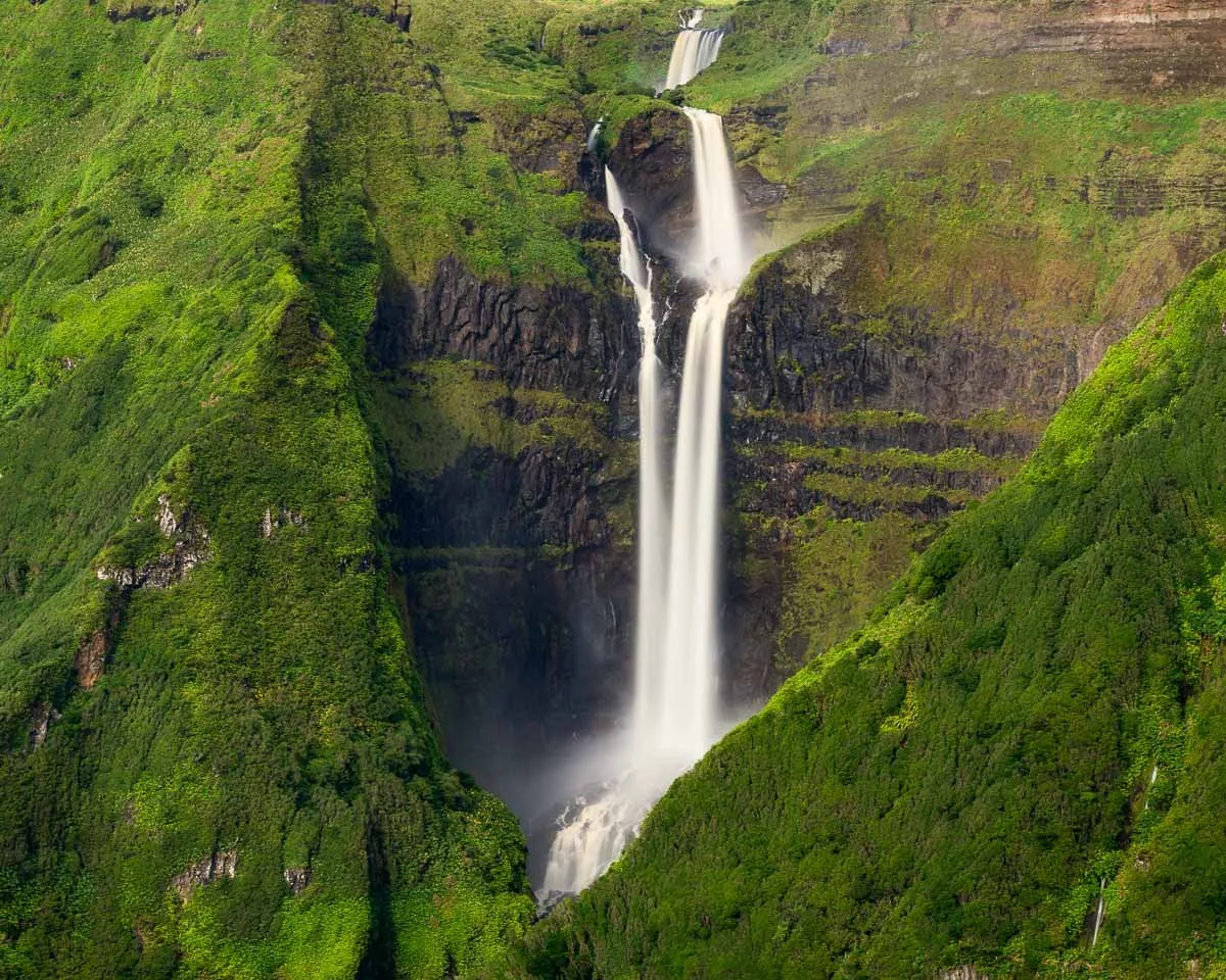 A tall waterfall cascading down a lush green mountain surrounded by dense forests.